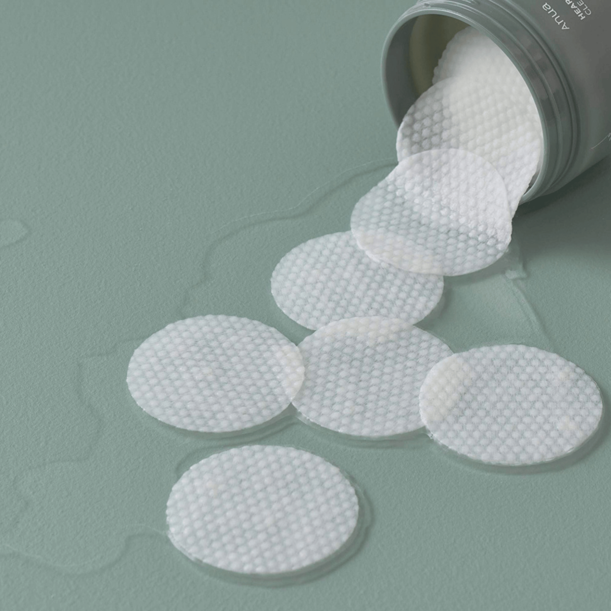 Nahaufnahme der strukturierten Anua Clear Pads, die aus der geöffneten Dose auf einen feuchten, grünen Untergrund fallen