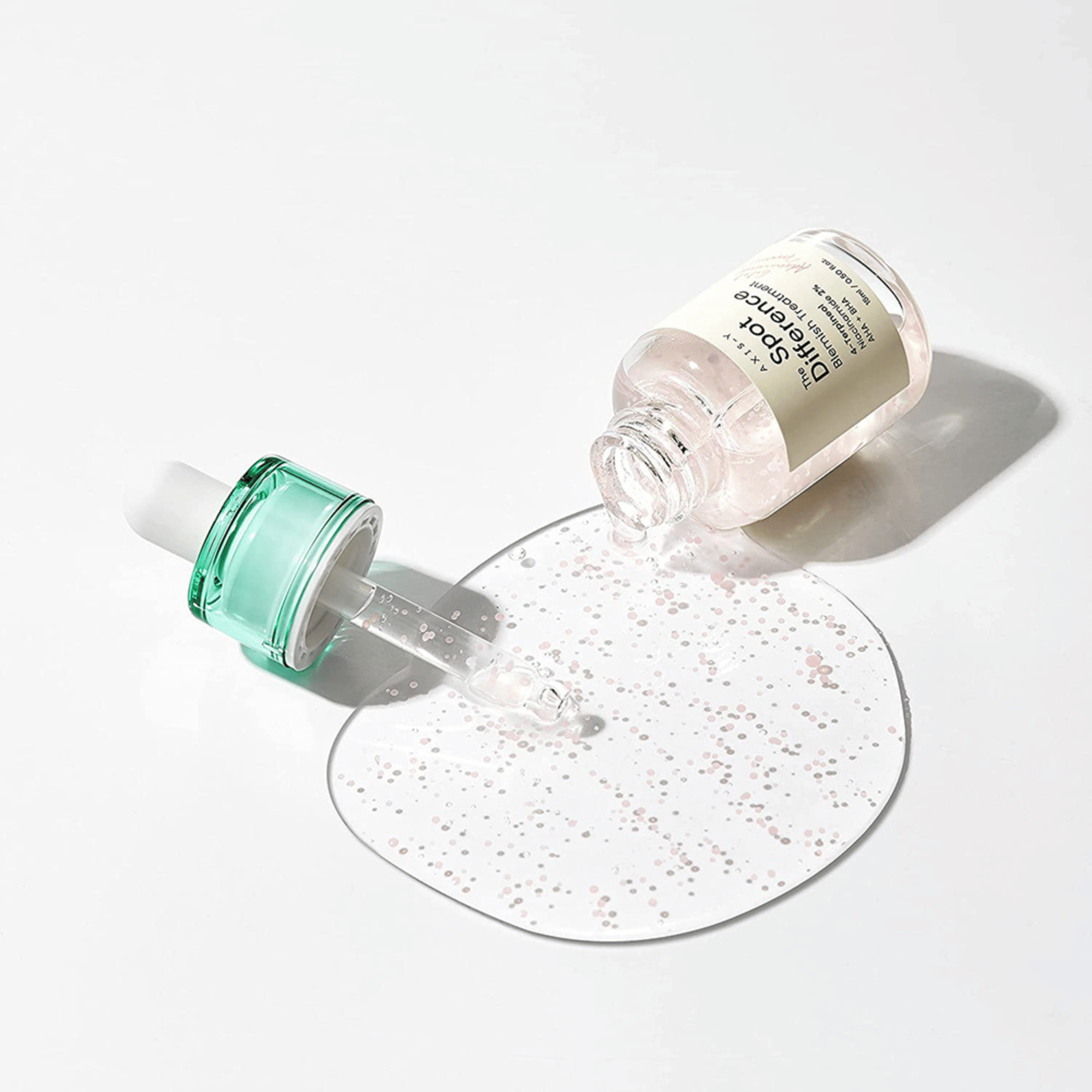 Offene Flasche des AXIS-Y Spot The Difference Blemish Treatment, daneben die Pipette mit transparenter Geltextur und pinken Perlen auf weißem Untergrund