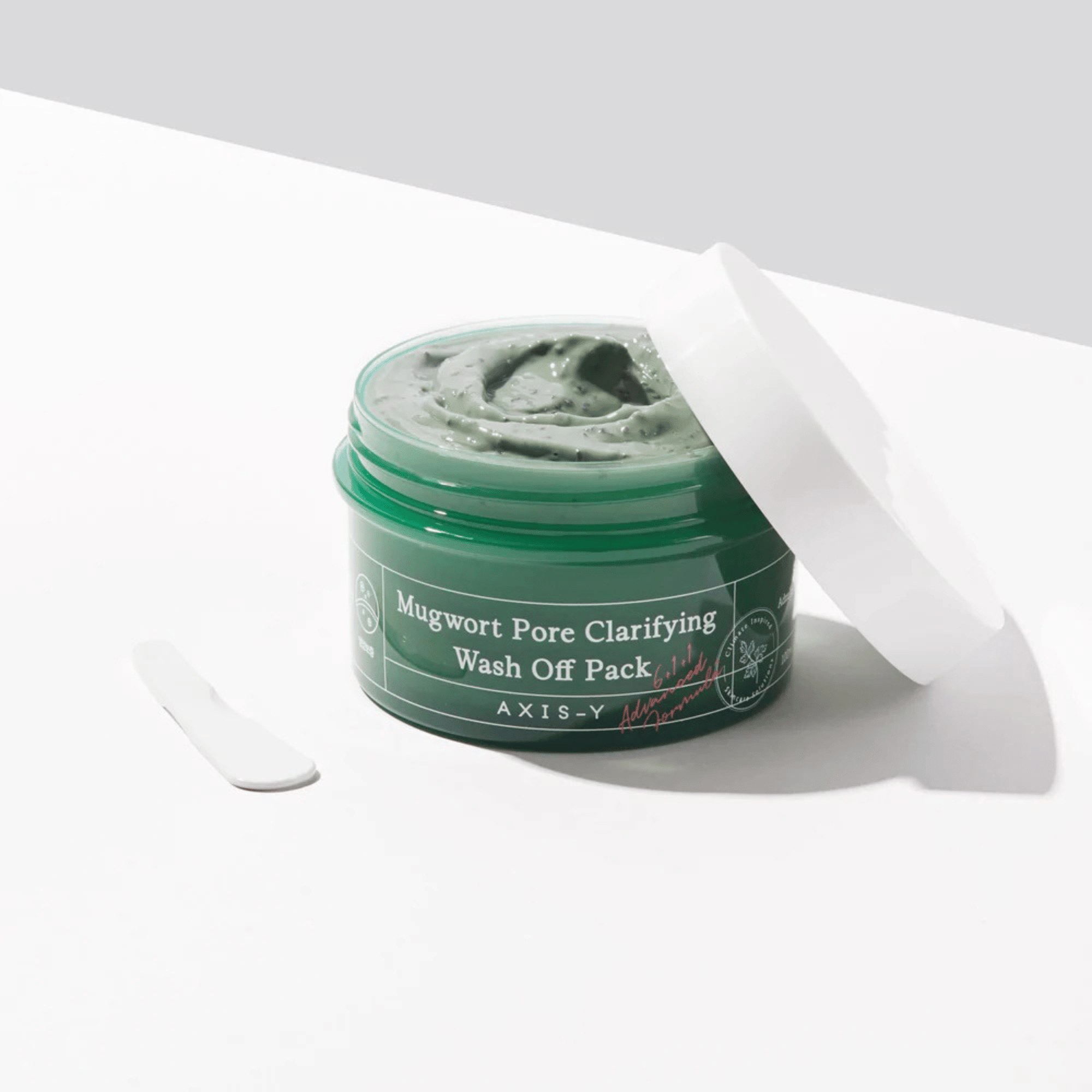 Offener Tiegel mit der Axis-Y Mugwort Pore Clarifying Wash Off Pack Maske, daneben ein Spatel auf weißem Untergrund