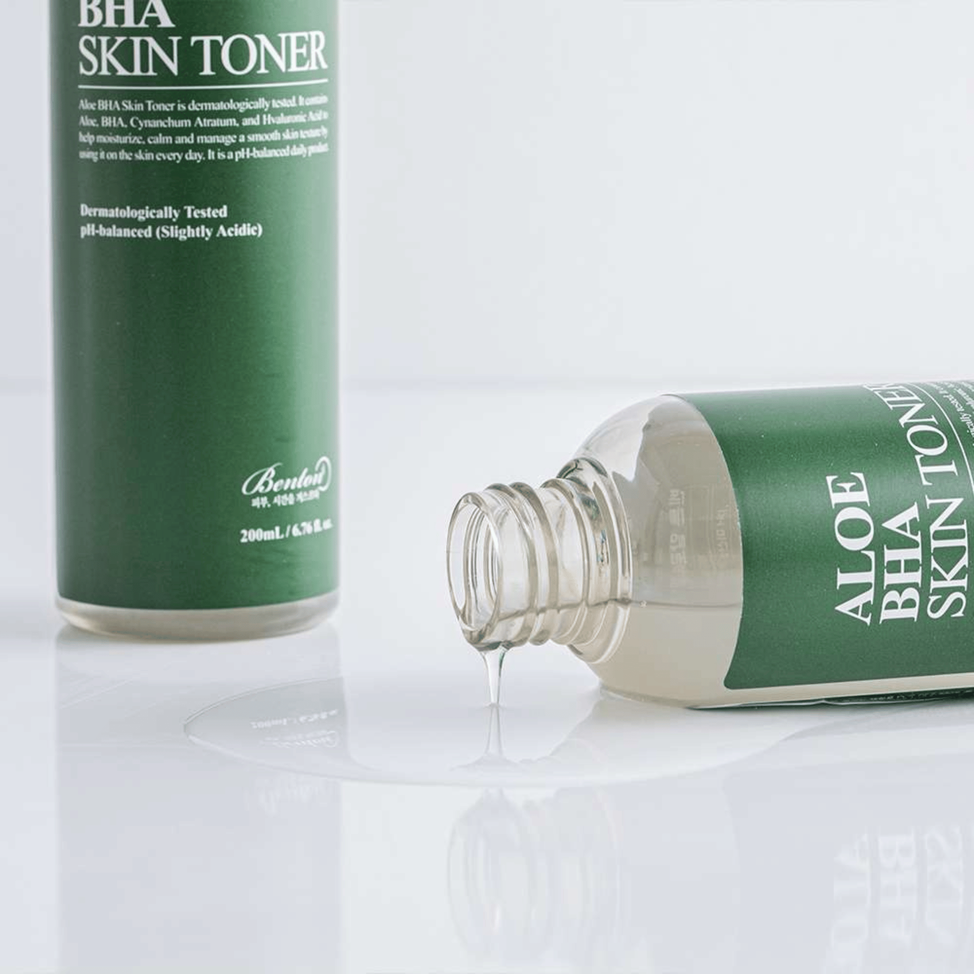 Aloe BHA Skin Toner liegt auf der Seite, wobei Toner auf eine weiße Oberfläche verschüttet ist, während eine andere aufrecht stehende Flasche teilweise im Hintergrund zu sehen ist.