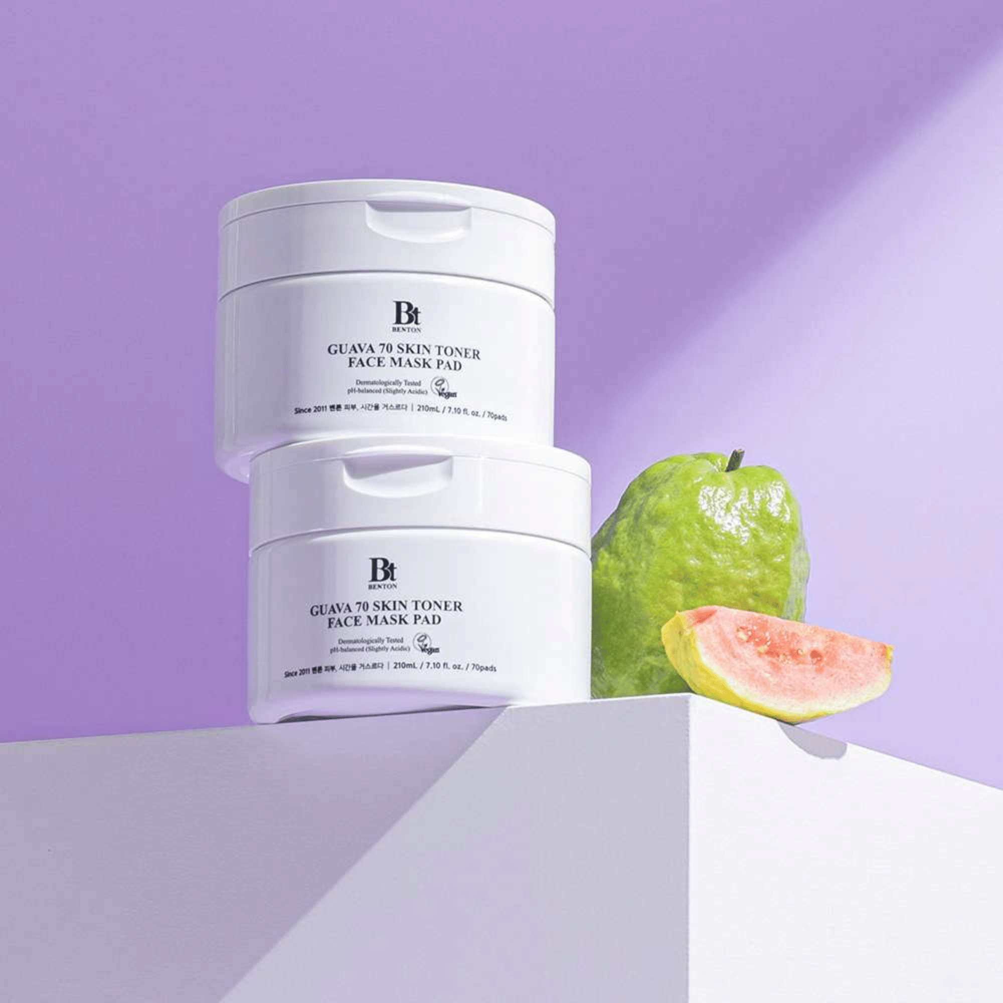 Zwei Benton Guava Skin Toner Face Mask Pad sind neben einer ganzen Guave und einer Scheibe auf einer weißen Fläche gestapelt, alles vor einem hellvioletten Hintergrund.