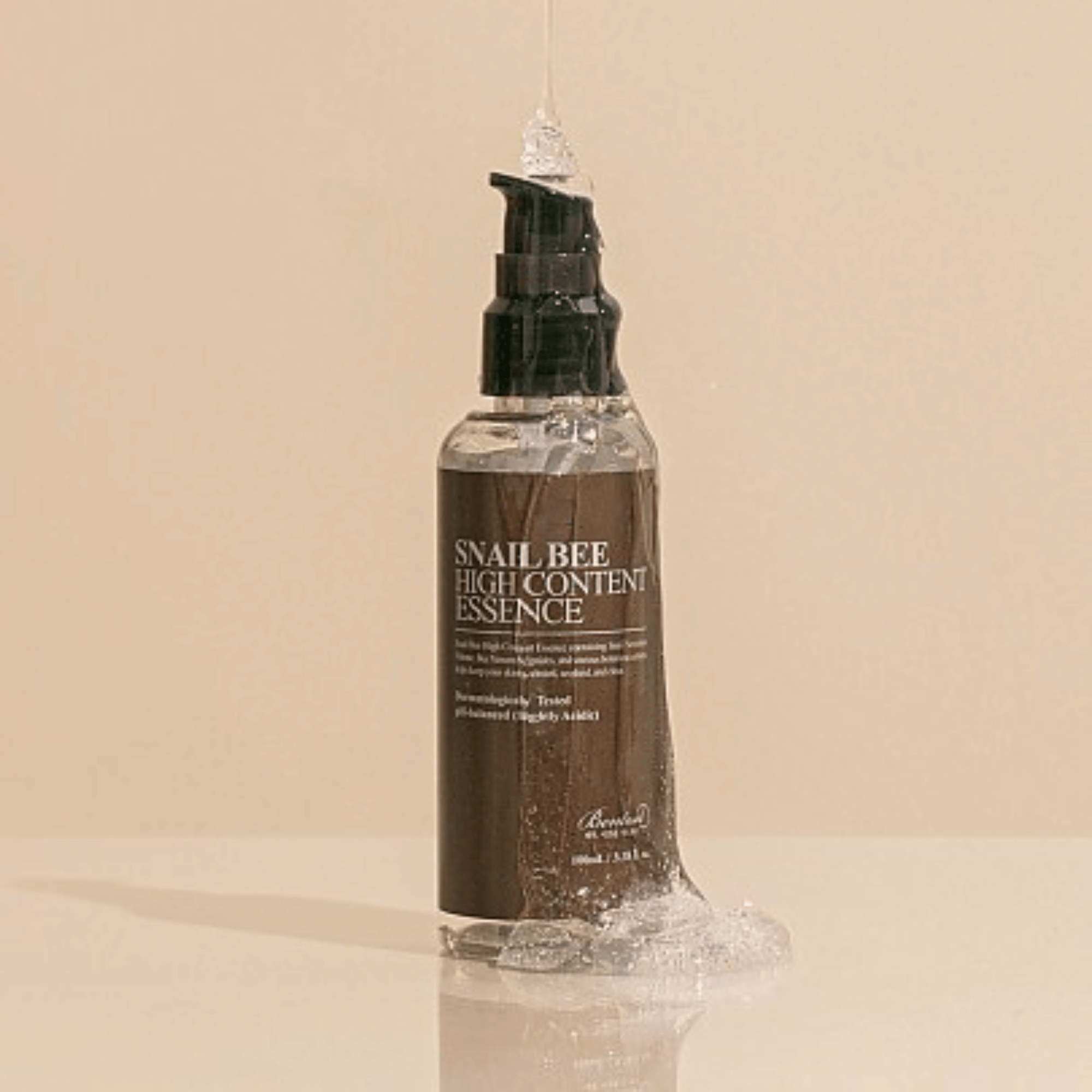 Eine Flasche Benton Snail Bee High Content Essence mit Wassertropfen darauf, präsentiert vor einem schlichten beigen Hintergrund.