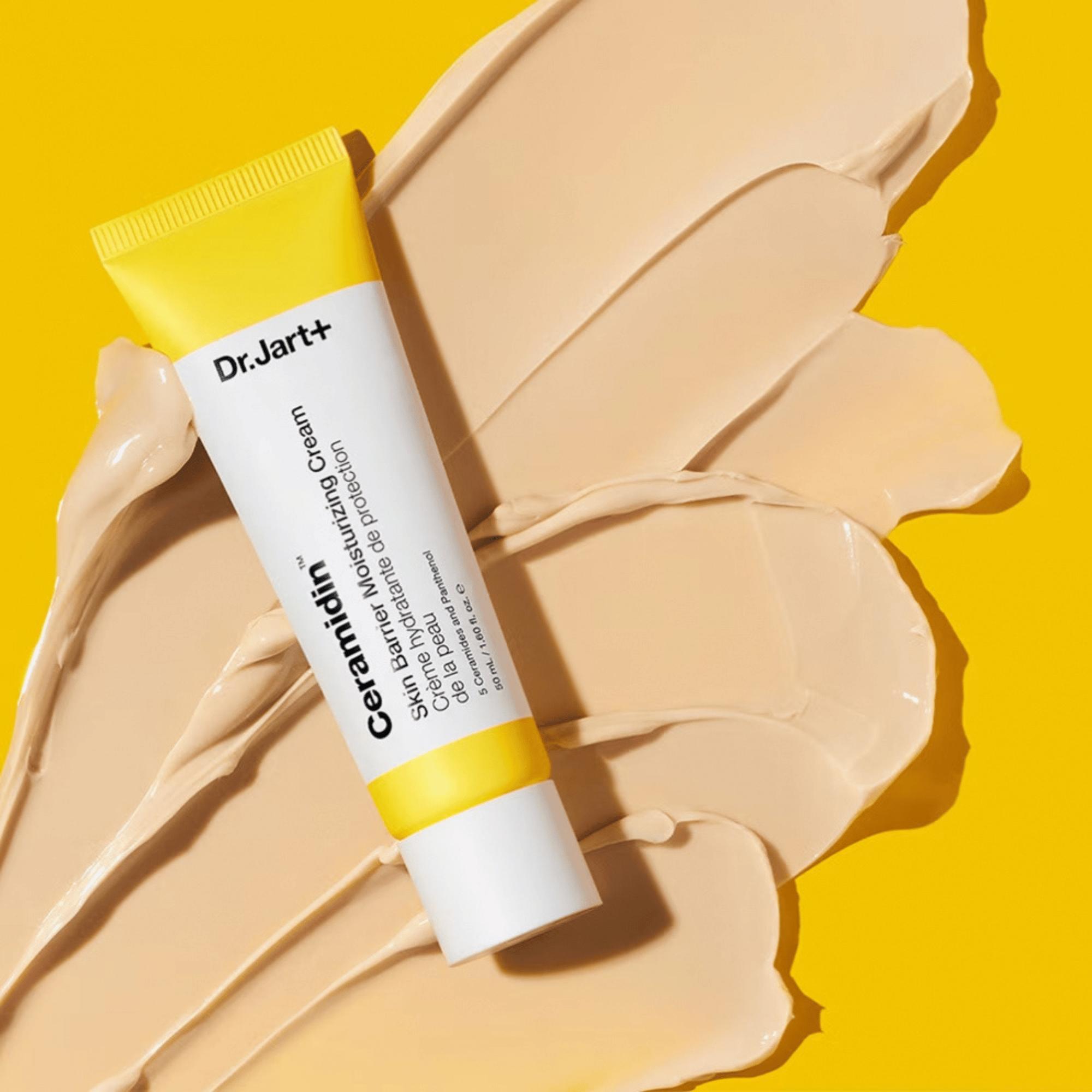 Eine Tube Dr. Jart+ Ceramidin Skin Barrier Moisturizing Cream steht auf beigen Cremeflecken vor einem leuchtend gelben Hintergrund.