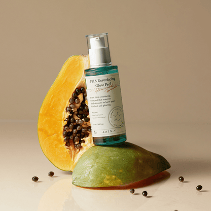 Axis-Y PHA Resurfacing Glow Peel steht auf halber Papaya mit aufgestellter Flasche, frische Kerne rundherum verstreut