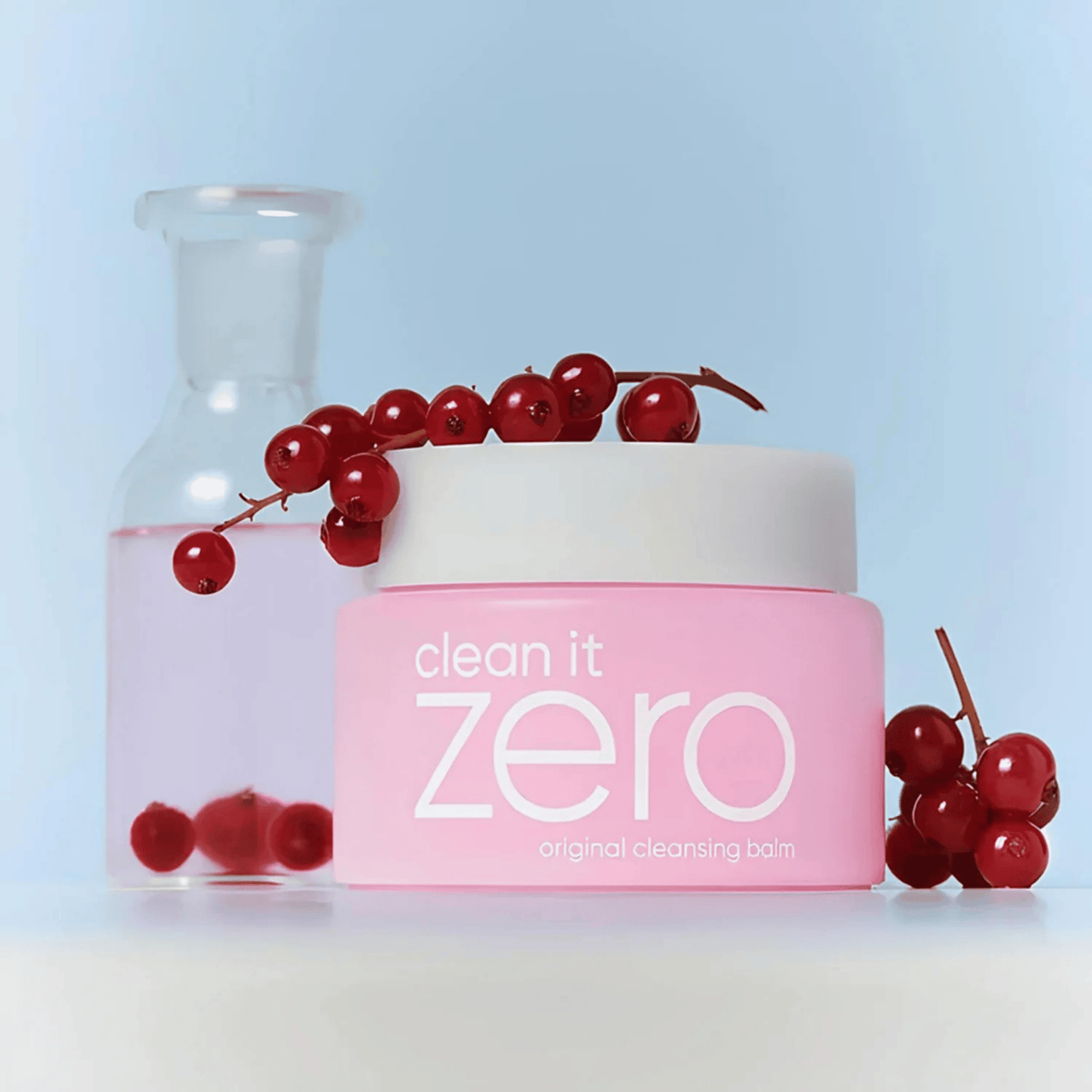 Ein rosafarbener Tiegel BANILA CO Clean It Zero Cleansing Balm Original 180ml wird zusammen mit roten Johannisbeeren und einer klaren Glasflasche, gefüllt mit rosafarbener Flüssigkeit und roten Johannisbeeren, präsentiert.