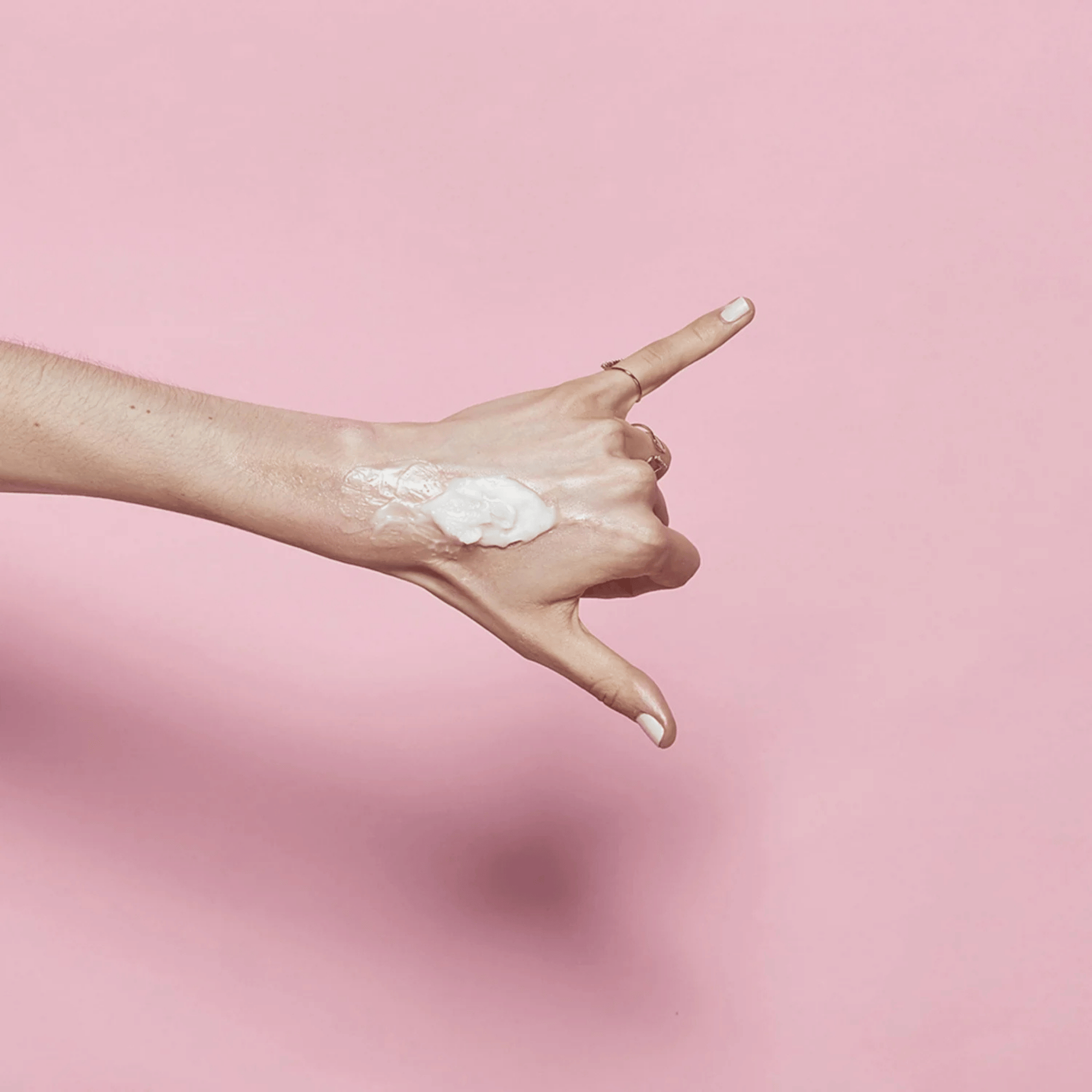 Eine Hand mit heller Haut macht ein &quot;Shaka&quot;-Zeichen vor einem rosafarbenen Hintergrund, auf dem BANILA CO Clean It Zero Cleansing Balm Original 180ml als kleine Menge weißer Creme auf dem Handrücken verteilt ist.